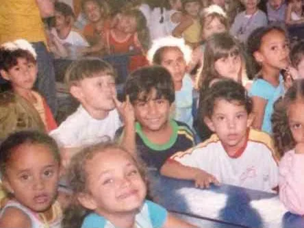 Elias criança, sentado entre colegas numa sala de aula cheia, fazendo o sinal de rock com os dedos.