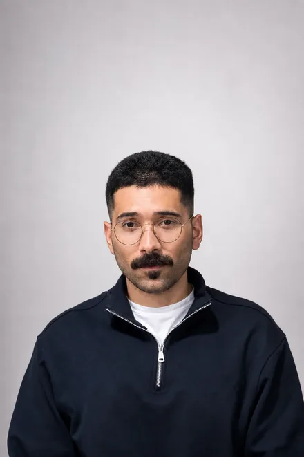 Retrato de Elias de frente para a câmera, de óculos redondos e moletom de gola com zíper, sobre fundo cinza claro.